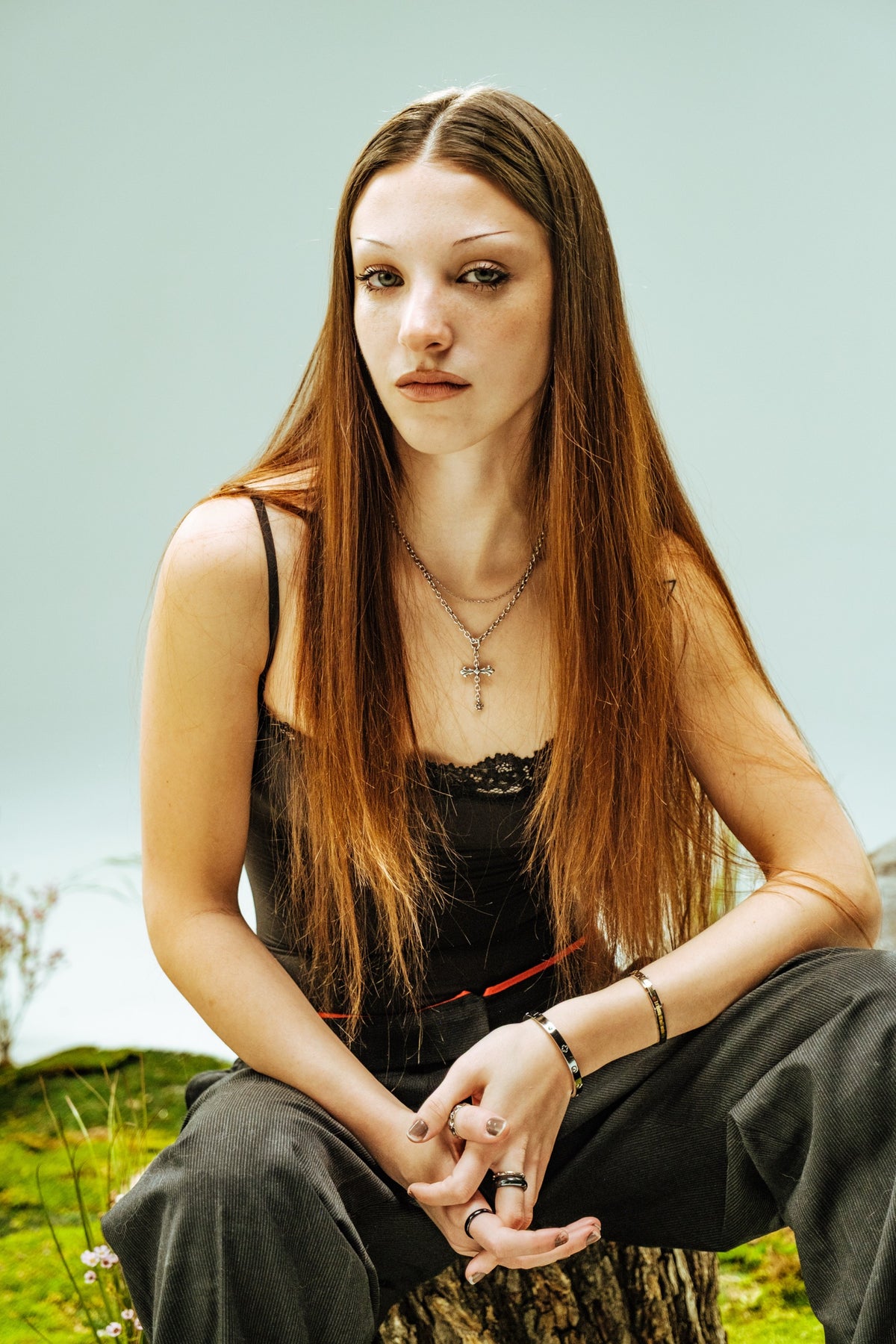A woman with long auburn hair sits on a tree stump on a moss set, hands clasped, looking steadily at the camera