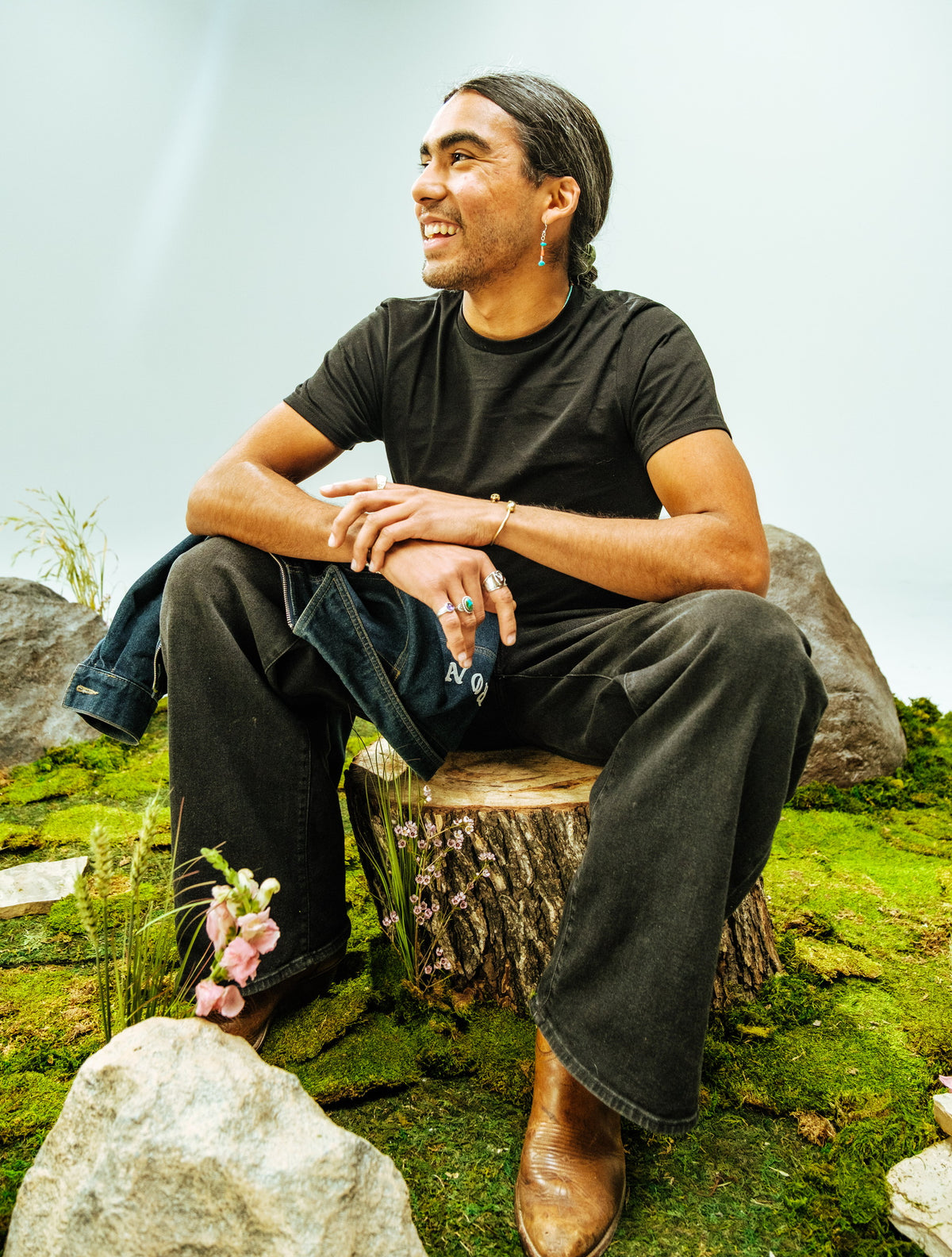 A man sits on a tree stump on a moss covered set, a denim jacket over one knee, smiling and looking off to one side