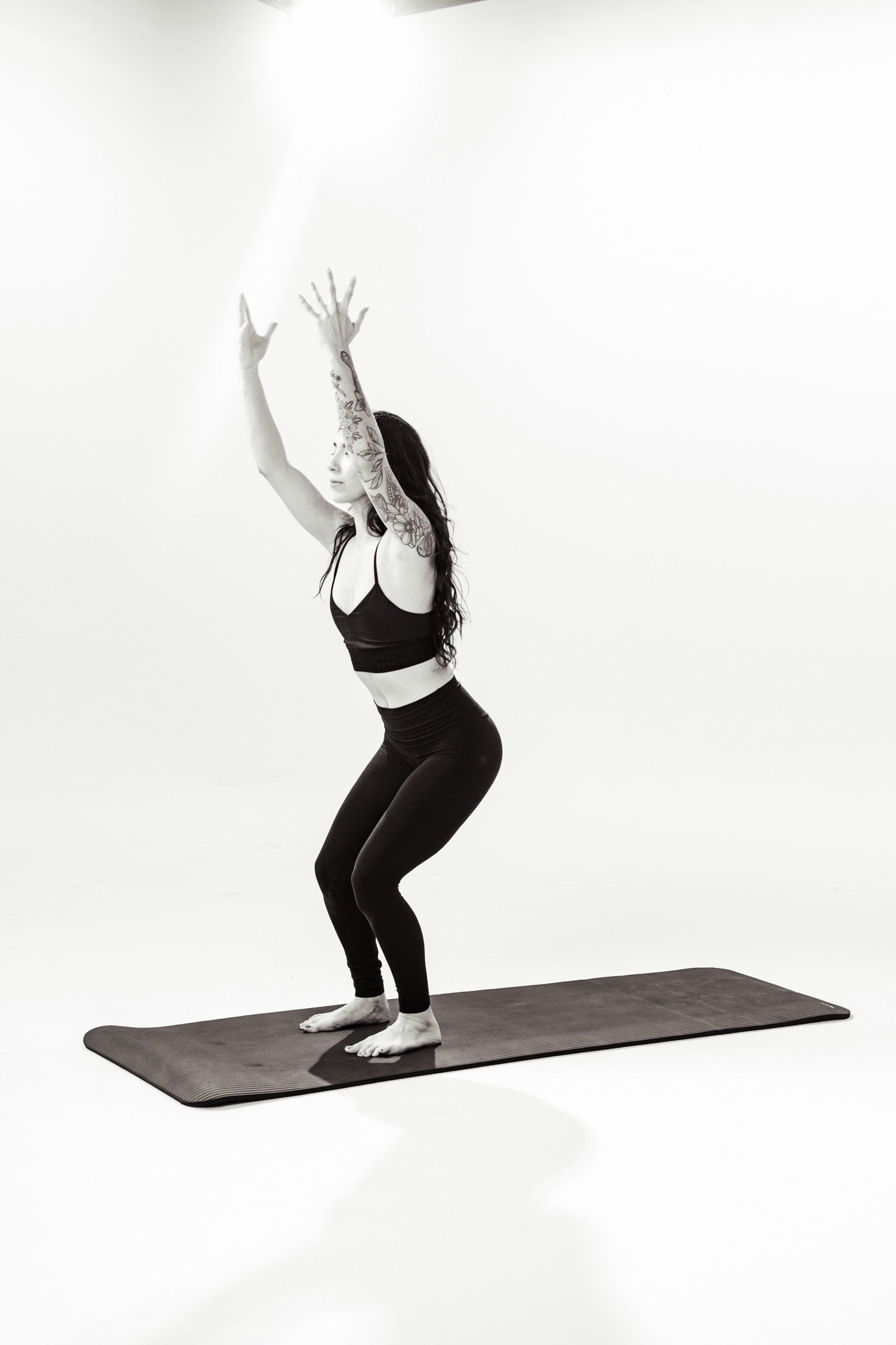 A woman in a deep squat with both arms raised overhead on a mat against a white studio backdrop