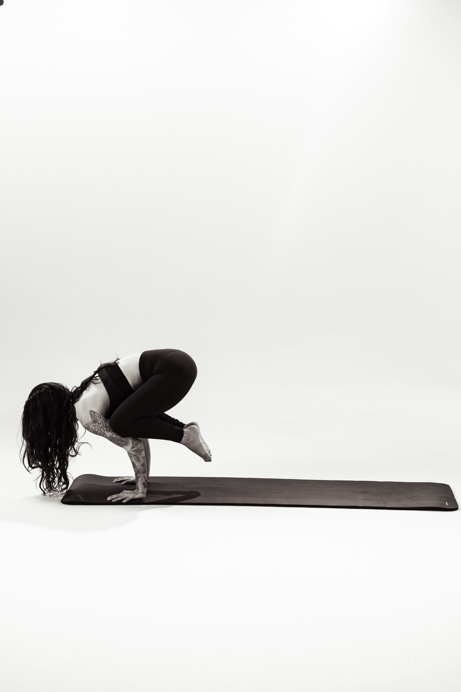 A woman balancing in a crow pose on a mat against a white studio backdrop