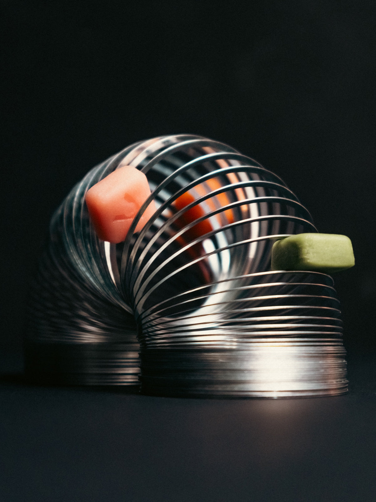 A metal spring toy on a dark tabletop with a pink gummy and a green gummy resting on its coils