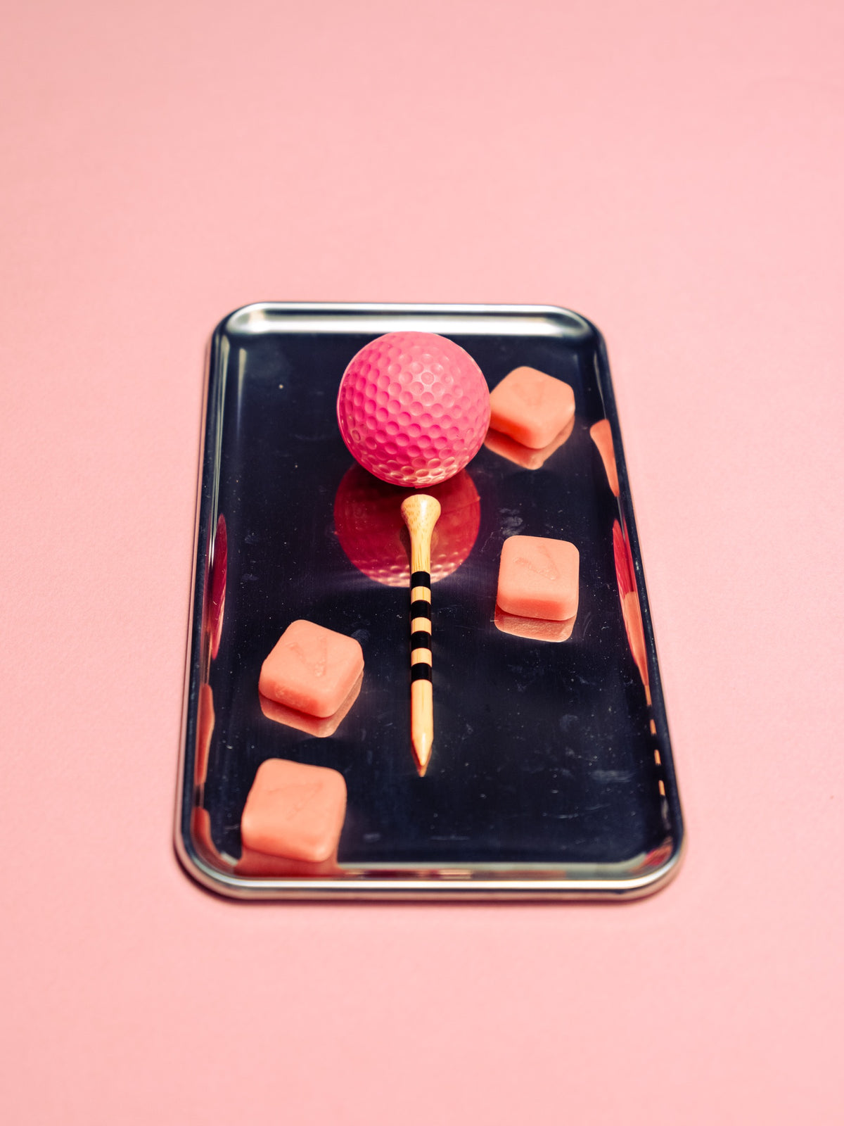 Pink gummies and a pink golf ball on a tee arranged on a steel tray against a pink backdrop