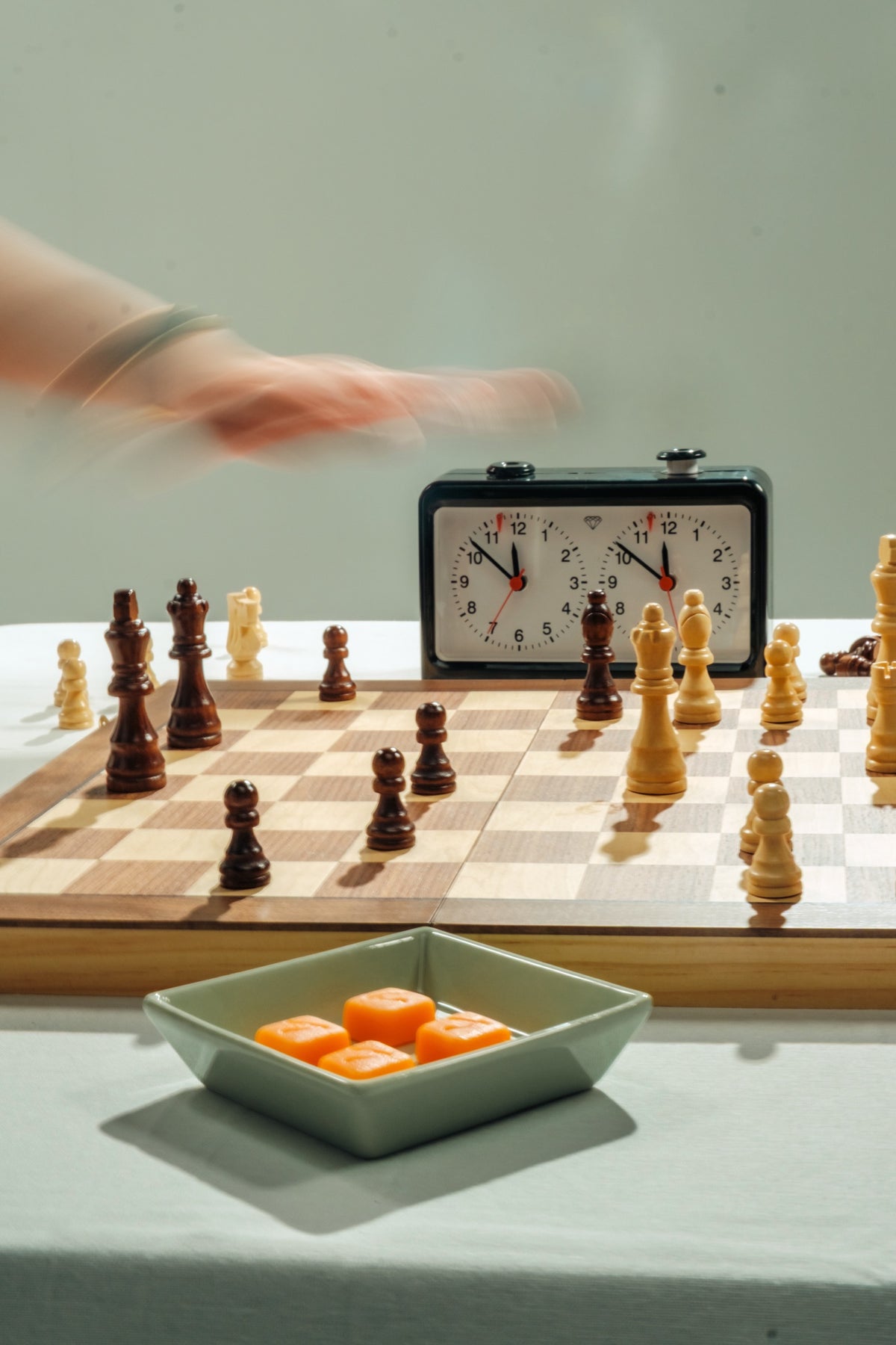 A hand in motion over a chessboard beside a game clock, with a dish of gummies on the board