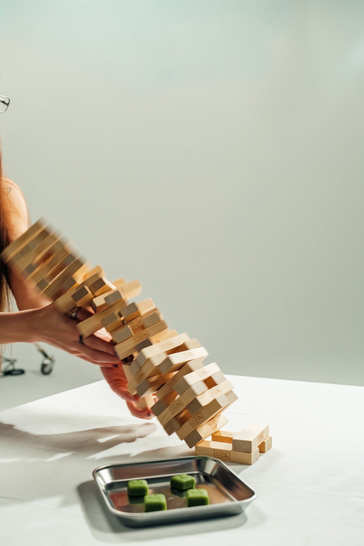 A hand pulling a block from a toppling wooden tower on a white table, with a tray of green gummies beside it